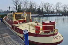 Classic 1927 Dutch Barge moored on a serene river, surrounded by trees and other boats.